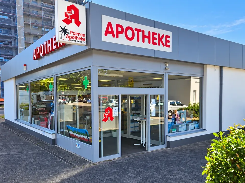Die Palmen Apotheke-Brückenhof an der Heinrich-Plett-Straße in Kassel-Oberzwehren: Eckeingang mit Schiebetür, roter Schriftzug „APOTHEKE" über der Fensterfront und das Logoschild an der Gebäudeecke.